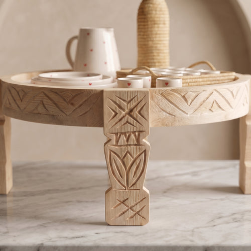 A round carved wooden serving tray table with decorative leg details, styled with white ceramic cups and a woven basket holder, set on a marble surface against a neutral beige background.
