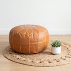 Caramel tan Moroccan leather pouf on a woven mat beside a small potted plant