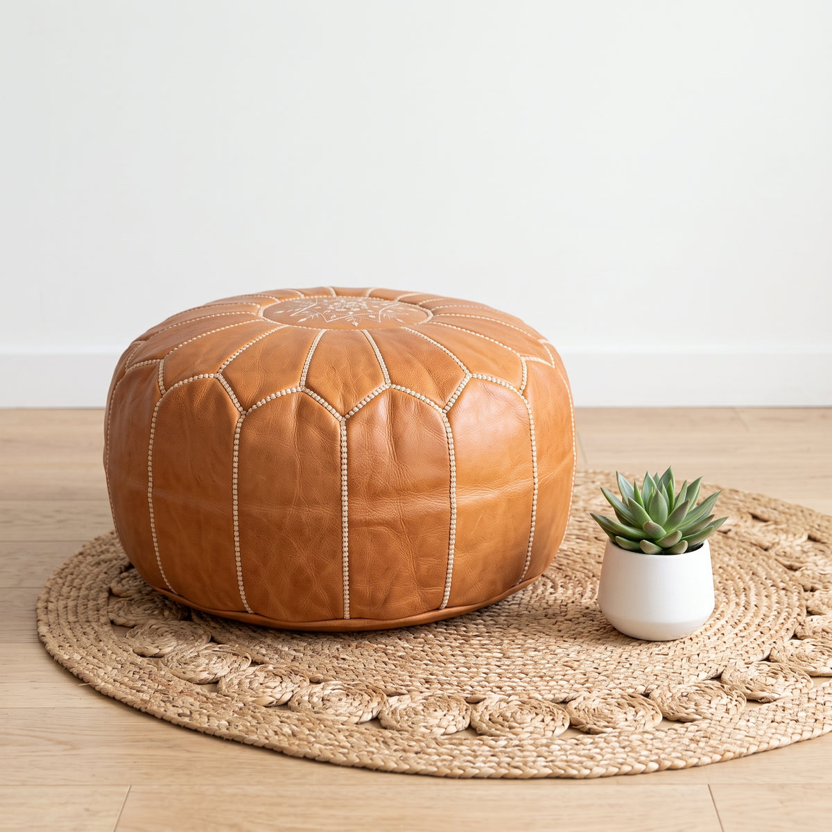 Caramel tan Moroccan leather pouf on a woven mat beside a small potted plant