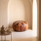 Two-tone brown leather pouf in a beige arched alcove beside a small brass table