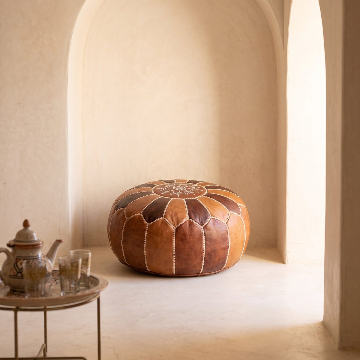 Two-tone brown leather pouf in a beige arched alcove beside a small brass table
