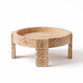 A round, low wooden tray table with carved geometric patterns on the rim and legs, featuring a central pedestal support, shown on a white background.
