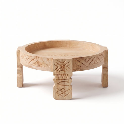 A round, low wooden tray table with carved geometric patterns on the rim and legs, featuring a central pedestal support, shown on a white background.