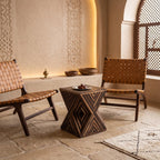 Dark wooden hourglass side table with geometric inlay beside woven wooden seating
