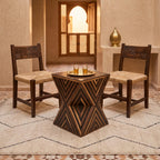 Dark geometric hourglass table topped with a brass tray between two woven-seat chairs