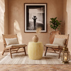 Yellow striped drum table with a vase between woven chairs beneath a framed photograph