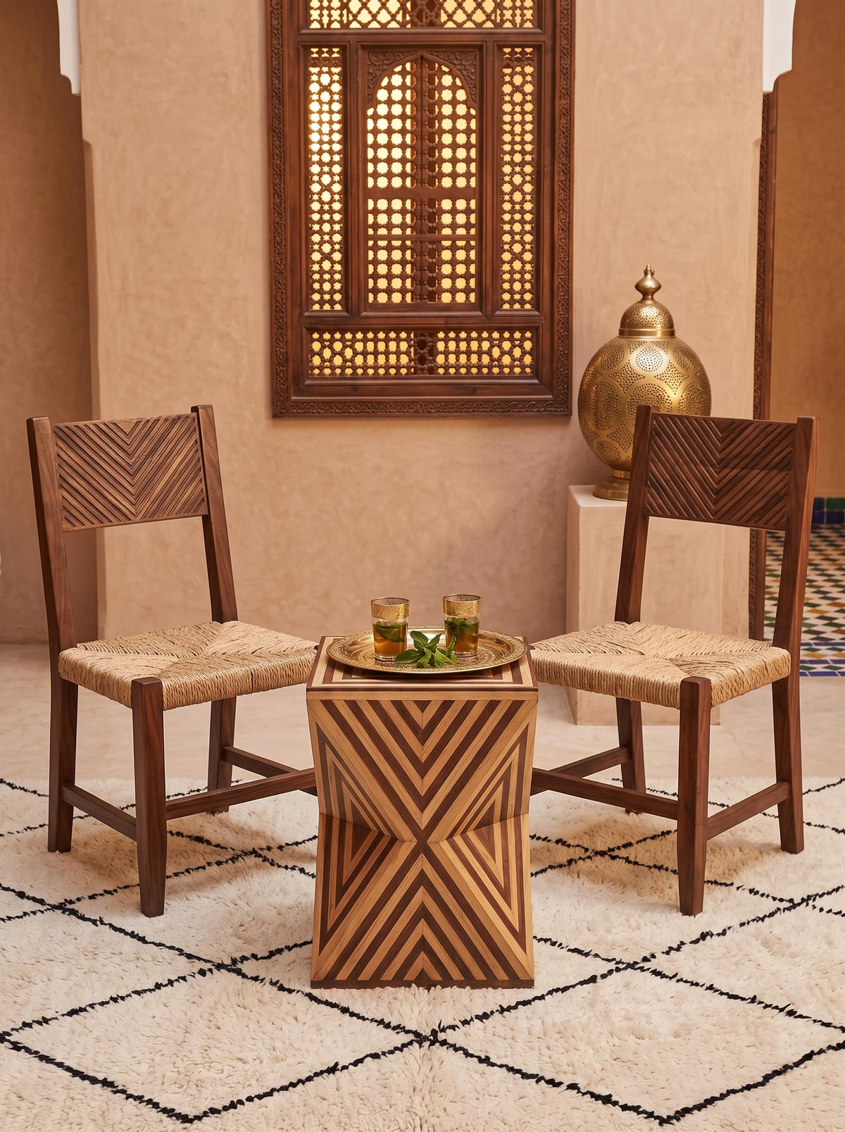 Chevron-patterned hourglass side table between two wooden chairs with a brass tea tray