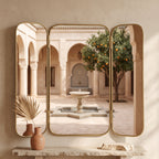 Gold brass triptych wall mirror above a stone ledge, reflecting a Moroccan courtyard with arches and an orange tree